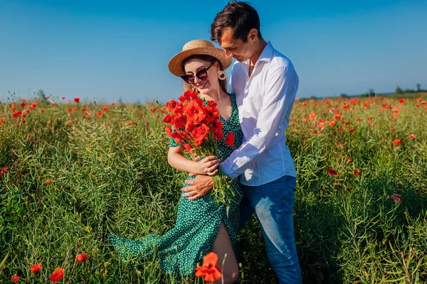 Любящая пара, гуляющая по маковому полю с букетом цветов. Счастливые мужчина и женщина обнимаются, улыбаясь. Летний отдых — стоковое фото