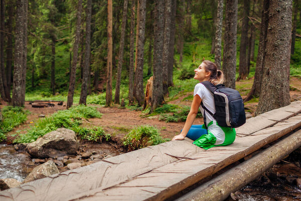 Traveler hiker with backpack sitting on bridge across river in Carpathian forest resting. Trip to summer Ukraine