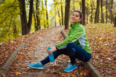 Koşucu sonbahar parkında antrenmandan sonra dinleniyor. Patikada elinde su şişesi tutan mutlu kadın. Sportif, aktif, sağlıklı yaşam tarzı. Temiz havada sonbahar egzersizi.