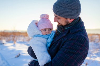 Baba kışın bebekle birlikte dışarıda yürüyor. Küçük kızı tutan adam sıcak kıyafetler giyiyor. Karların arasında aile kucaklaşması güneşli manzaranın tadını çıkarıyor