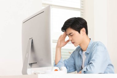 young Asian man with headache, sitting in front of computer, workover concept clipart