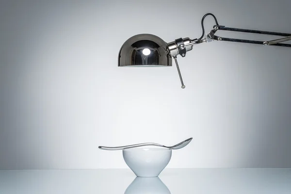 Verlichting van witte ceramische kom zilveren lepel met bureaulamp — Stockfoto