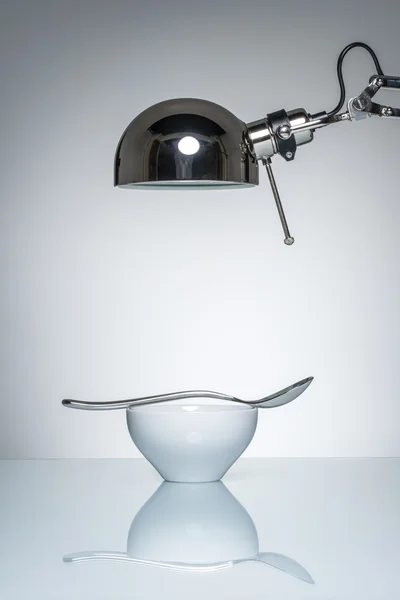Verlichting van witte ceramische kom zilveren lepel met bureaulamp — Stockfoto