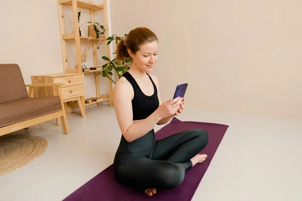 Yoga yaptıktan sonra spor yapan genç bir kadın, egzersiz yapmaya ara vermek, yoga minderinde dinlenmek, cep telefonuyla mesajlaşmak, akıllı telefon tutmak, arkadaşça gülümseyen mesajlar, kapalı kapılar, yoga stüdyosu.