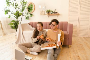 Genç erkek ve kadın çift evde dizüstü bilgisayarla pizza yiyorlar..