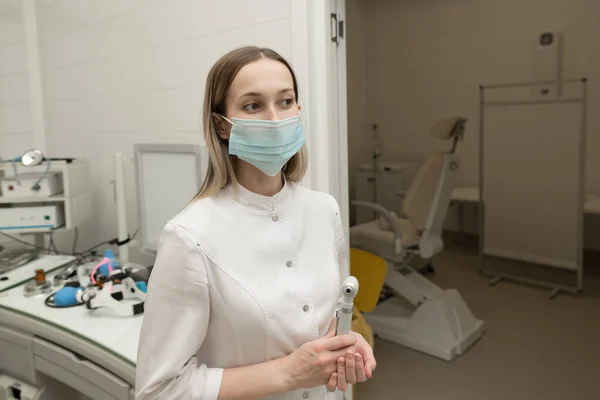 Hastanenin modern ENT ofisinde bir KBB doktoru elinde bir otoskop ile beyaz önlük ve maskeyle kameraya bakar.
