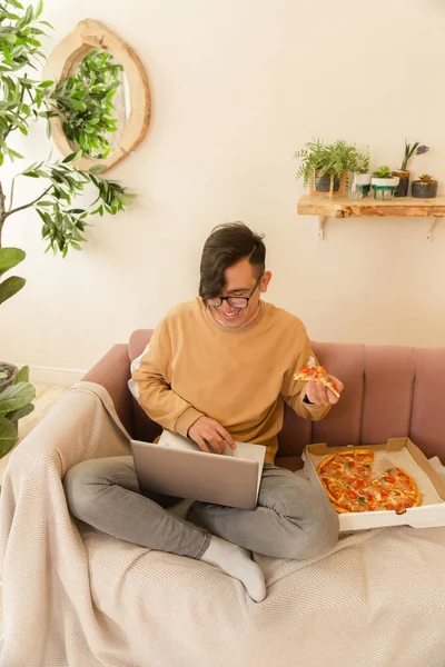 Evde dizüstü bilgisayarla pizza yiyen genç ve mutlu bir öğrenci.