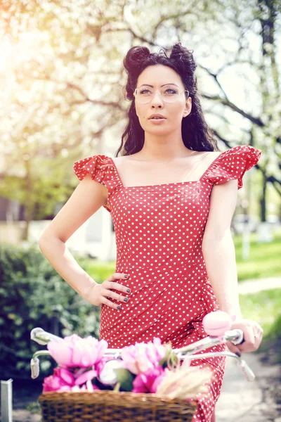 Donna sexy che indossa un abito vintage. pin-up seduta in bicicletta con alcuni fiori colorati nel cestino nel centro storico — Foto Stock