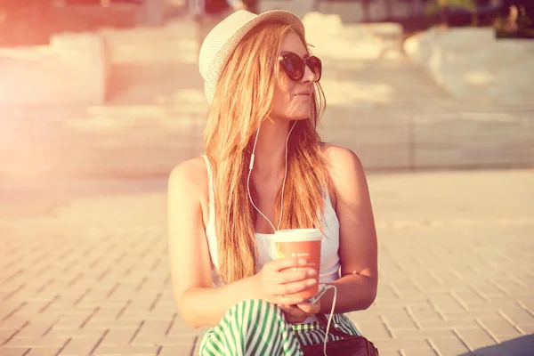 Retrato de una niña en un sombrero caminando en un parque de la ciudad con una taza de café de papel. Disparos al atardecer . — Foto de Stock