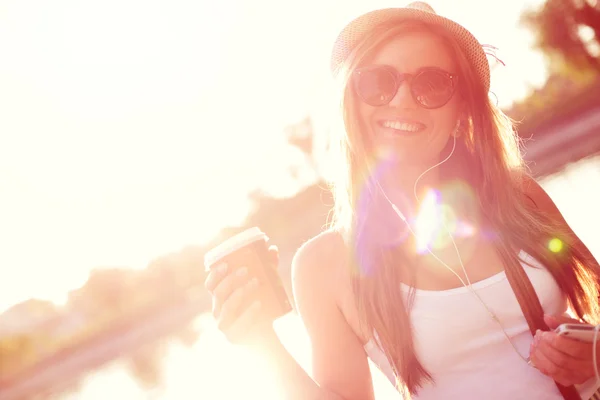 Porträt eines Mädchens mit Hut, das in einem Stadtpark mit einem Kaffee aus Papier spaziert. Schießen bei Sonnenuntergang. — Stockfoto