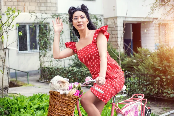 Sexy Frau im Vintage-Kleid. Pin-up sitzt auf Fahrrad mit ein paar bunten Blumen und einem kleinen Hund im Korb in der Altstadt — Stockfoto