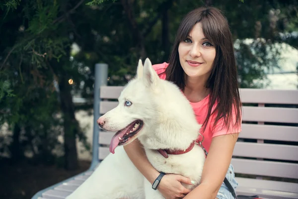 Chica joven sentada en el banco y jugando con su perro blanco husky —  Fotos de Stock