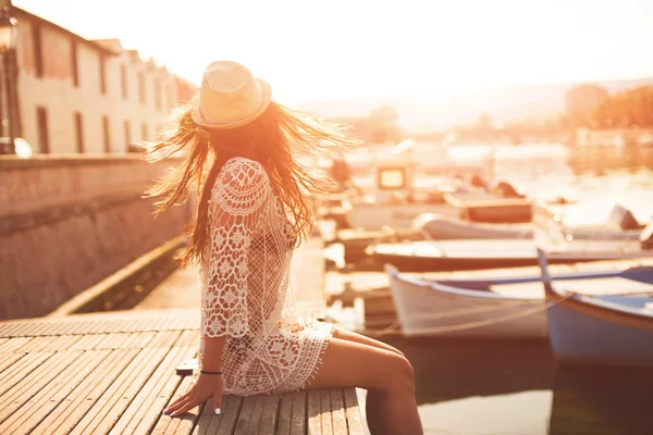 Ung kvinna i hatt och söt sommarklänning står på bryggan med fridfull stad landskap, tittar på solnedgången — Stockfoto