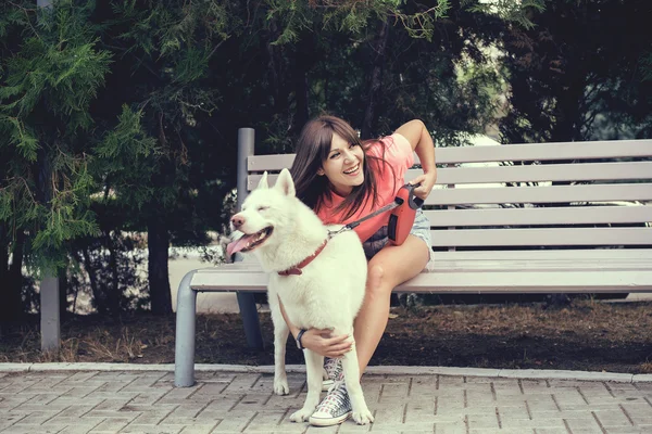 Chica joven sentada en el banco y jugando con su perro blanco husky —  Fotos de Stock