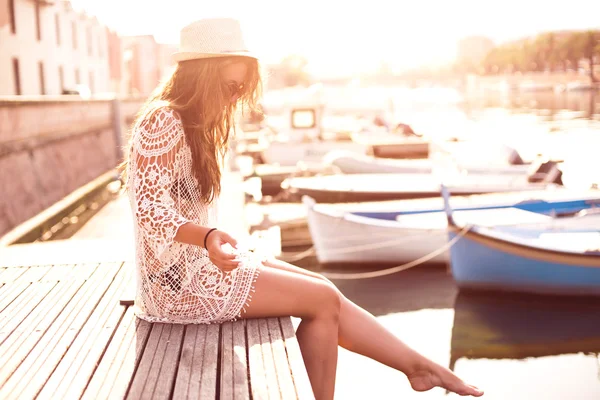 Ung kvinna i hatt och söt sommarklänning står på bryggan med fridfull stad landskap, tittar på solnedgången — Stockfoto