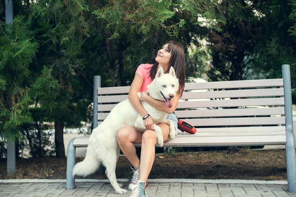 Jovem sentada no banco e brincando com seu cão branco husky — Fotografia de Stock