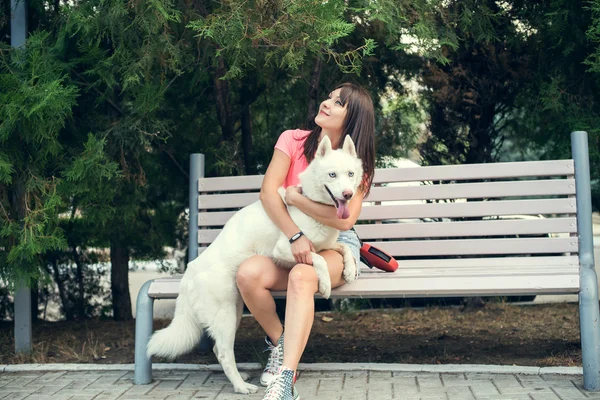 Chica joven sentada en el banco y jugando con su perro blanco husky —  Fotos de Stock