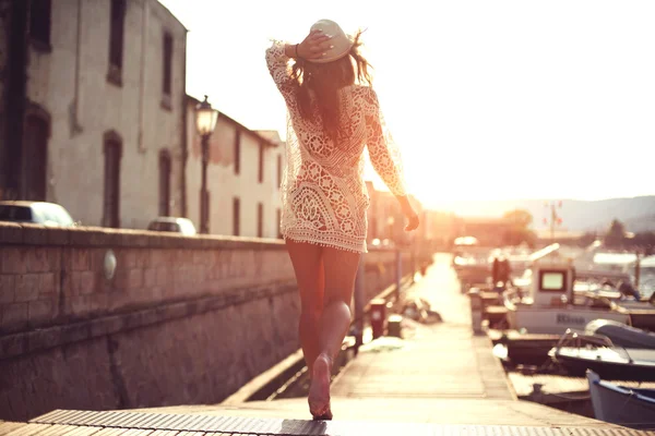 Young woman in hat and cute summer dress standing on the pier with peaceful town scenery, looking at sunset