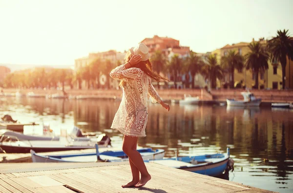 Mladá žena v klobouku a roztomilý letní šaty stojí na molu s poklidného městečka scenérie, při pohledu na západ slunce — Stock fotografie
