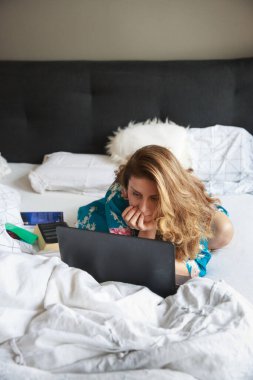 Gecelik giymiş güzel bir kadın, yatakta uzanıyor, bilgisayarla çalışıyor. Ev kavramından çalış.