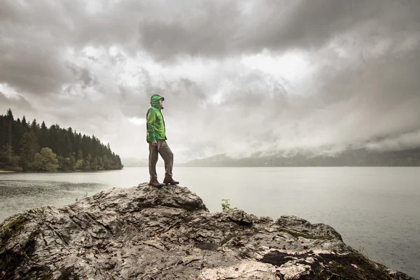 Mann steht im Regen auf einem Felsen neben einem Bergsee — Stockfoto