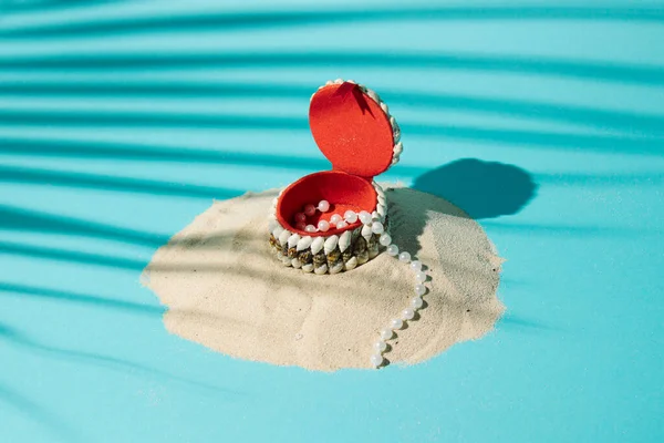 Beach sand with a box of shells and with pearls coming out of the box on a blue background with palm leaf shadow. Summer minimal flat lay beach concept.