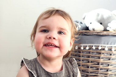 İki yıllık gülümseyen mutlu kız, arka planında gri gözleri olan oyuncak dolu bir sepet, yakın plan.