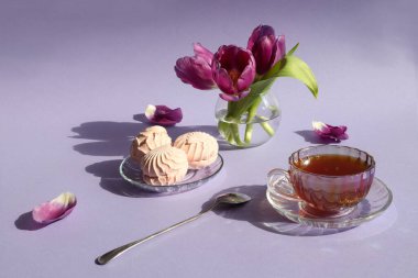 Bir çay fincanı çay tabağında marşmelov, bir vazo dolusu lale, bir çay kaşığı güneşte pastel bir zemin ve nesnelerin gölgesi.