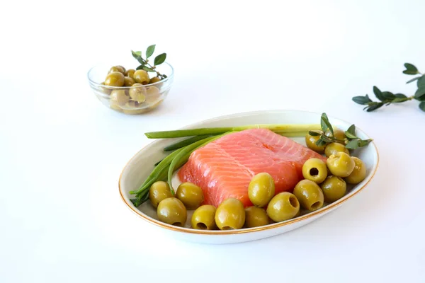 Red fish with olives, green onions on a platter on the background of a cup with olives on a white background, side view