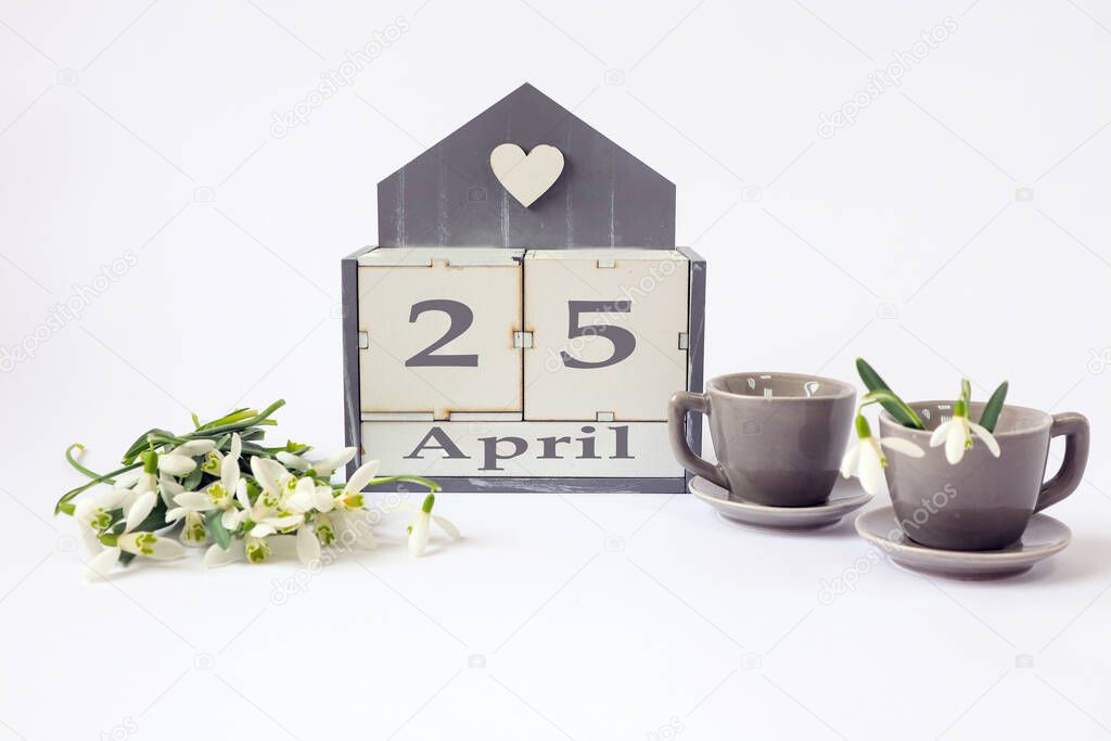 Calendar for April 25: cubes with the number 25, the name of the month in English, two gray coffee cups, a bouquet of snowdrops on a light background
