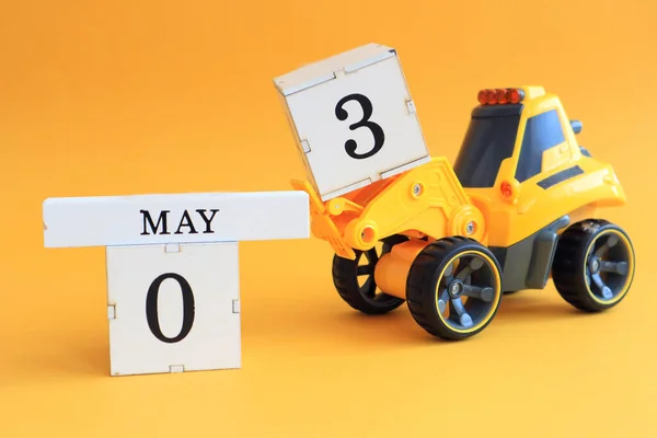 Calendar for May 3: a toy yellow tractor with numbers on cubes 0 and 3, the name of the month in English on a yellow background