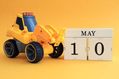 10 Mayıs takvimi: Küpte 10 numaralı oyuncak sarı traktör, sarı arka planda ayın İngilizce ismi.