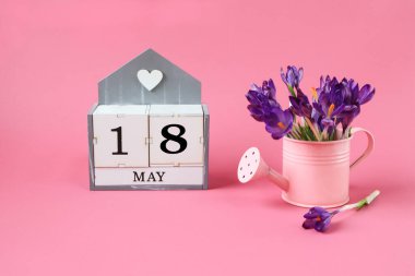 18 Mayıs takvimi: 18 numaralı bir küp, Mayıs ayının adı İngilizce, pembe arka planda bir buket mor timsah olan pembe bir sulama kutusu.