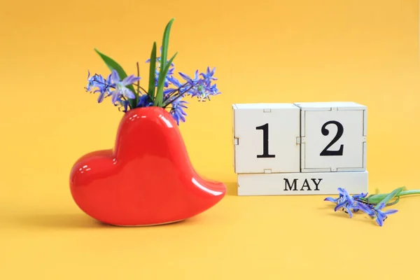 Calendar for May 12 : a bouquet in a heart-shaped vase with blue flowers and the numbers 12 on cubes, the name of the month of May in English, yellow background