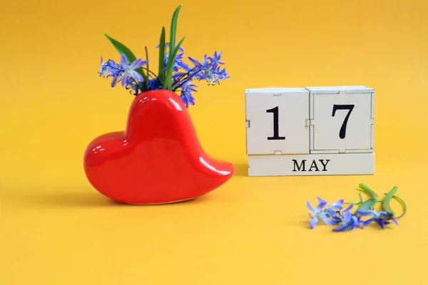 Calendar for May 17 : a bouquet in a heart-shaped vase with blue flowers and the numbers 17 on cubes, the name of the month of May in English, yellow background