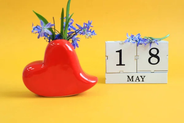 Calendar for May 18 : bouquet in a heart-shaped vase with blue flowers and the numbers 18 on cubes, the name of the month of May in English, yellow background