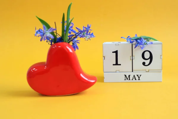 Calendar for May 19 : a bouquet in a heart-shaped vase with blue flowers and the numbers 19 on cubes, the name of the month of May in English, yellow background