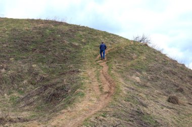 Mavi ceketli ve kot pantolonlu bir adam dik bir dağa tırmanır. Bahar manzaralı bir alanda yürür.