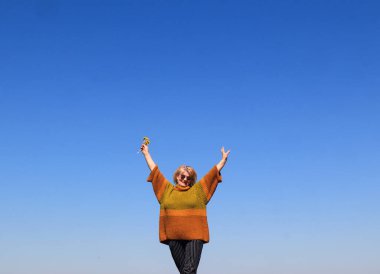 Mavi gökyüzüne karşı güneş gözlüklü yaşlı bir kadın ellerini havaya kaldırdı. Doğada hoş bahar yürüyüşleri kavramı.