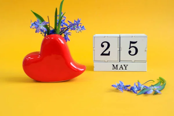 Calendar for May 25 : a bouquet in a heart-shaped vase with blue flowers and the number 25 on cubes, the name of the month of May in English, yellow background