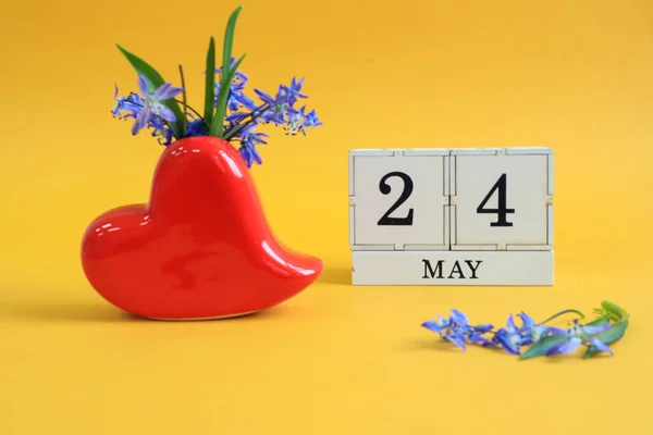 Calendar for May 24 : a bouquet in a heart-shaped vase with blue flowers and the number 24 on cubes, the name of the month of May in English, yellow background