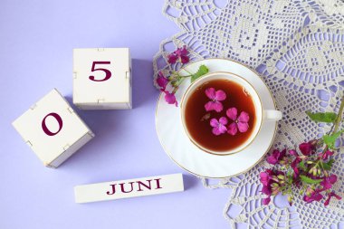5 Haziran için takvim: 0 ve 5 sayılı küpler, Haziran ayının ismi, içinde mor çiçekler olan bir fincan çay, açık arkaplanda gri bir açılış peçetesi, üst görünüm