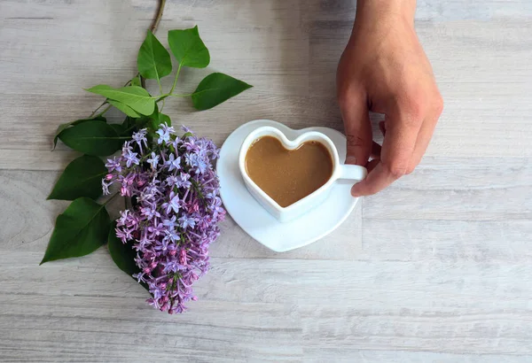 The concept of good morning and good mood.A cup of coffee in the shape of a heart in a man\'s hand, a branch of lilac on a gray background, a top view, a place for text .