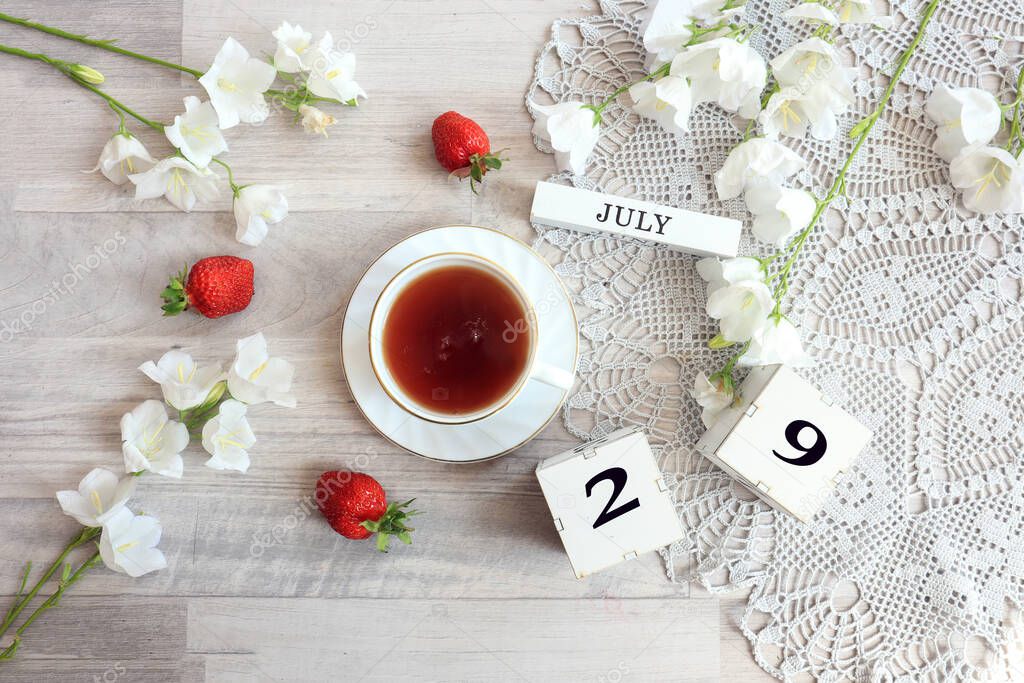 Calendar for July 29 : cubes with the number 29, the name of the month of July in English, a cup of tea on a gray openwork napkin, scattered flowers of bluebells and strawberries, top view