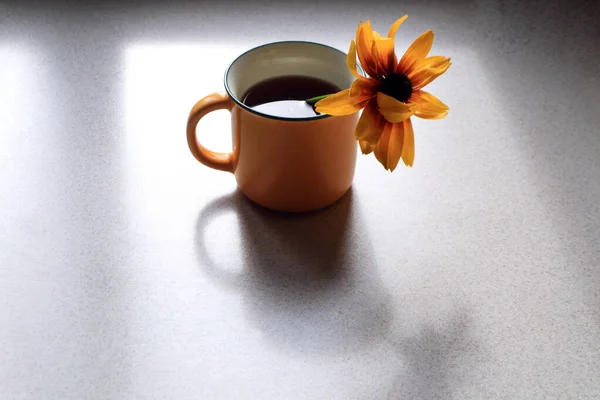 The concept of a good evening. A yellow mug of tea with a yellow flower on a gray kitchen table with shadows from objects, a place for text