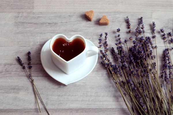 The concept of pleasant memories over a cup of tea. A cup of tea in the shape of a heart, two pieces of sugar, a bouquet of lavender on a gray background, side view, space for text