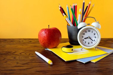Eğitim kurumlarında öğretmenlik kavramı. Bir saat, okul defterleri, kırmızı bir elma, kalemler, ahşap bir masa, sarı bir arka plan, bir kenar görüş, bir metin yeri. .