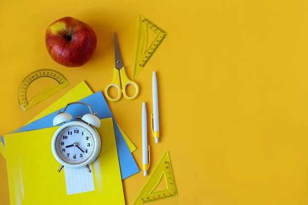 The concept of teaching in educational institutions. A red apple, school supplies-notebooks, pens, a stapler, a sharpener-a watch , scissors on a yellow background, a top view, a place for text.