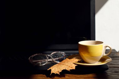 İyi bir sonbahar sabahı ve iş gününün başlangıcı kavramı. Sabah güneşi altında bilgisayar klavyesinde sarı bir fincan çay, sonbahar akçaağaç yaprağı, görüş için gözlük, yan görüş