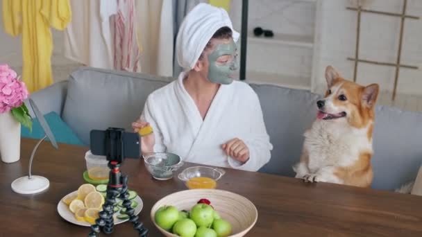 A Beauty Blogger in a Robe with a White Towel on Her Head Apply a Green Moisturizing Mask to Her Face and Blogs about Skin Care and Cosmetics. O Cão Bonito dela está sentado perto. Conceito de entretenimento — Vídeo de Stock